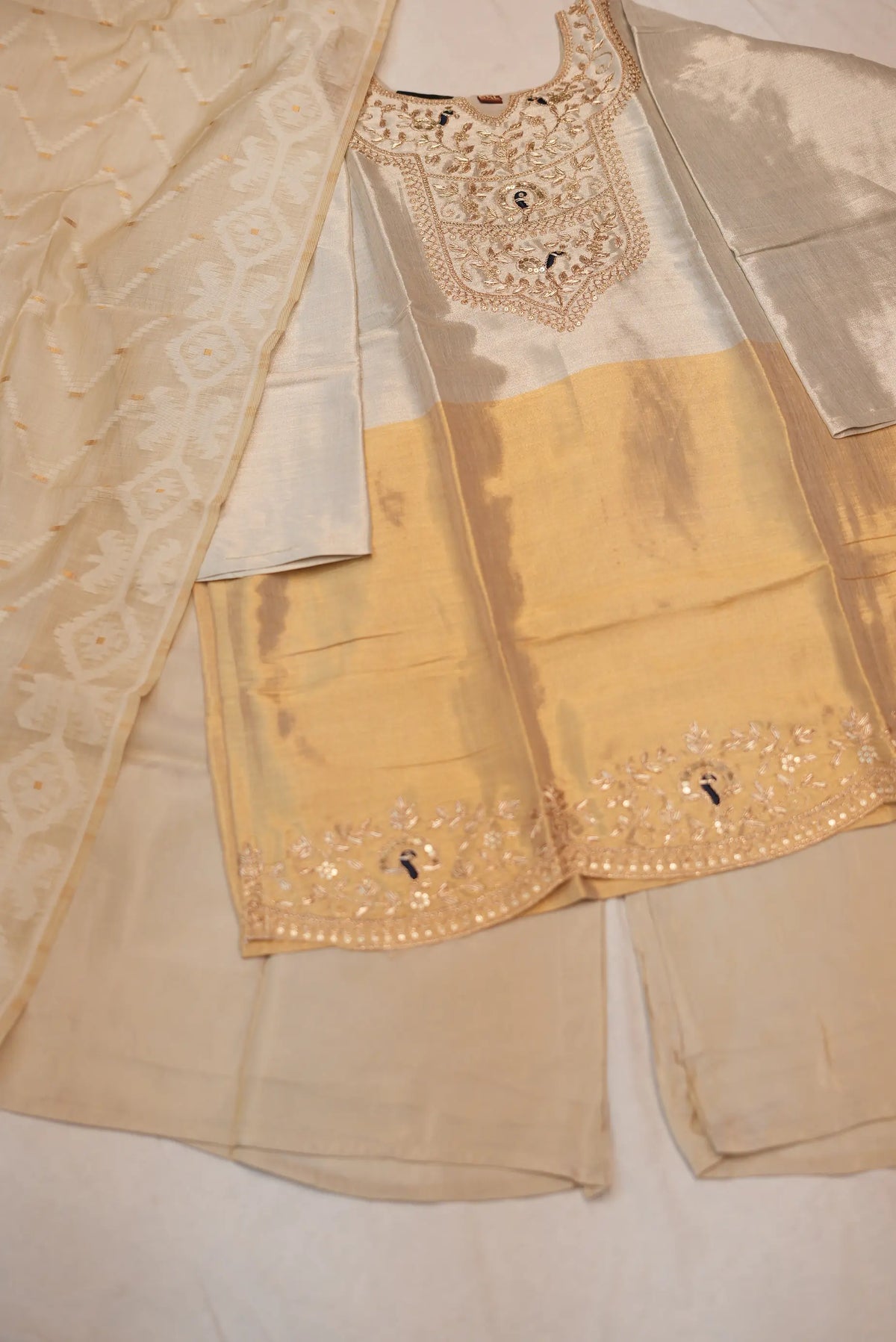 Flat lay view of 3 piece metallic off white and golden tissue short kurti suit set with palazzo and heavy dupatta